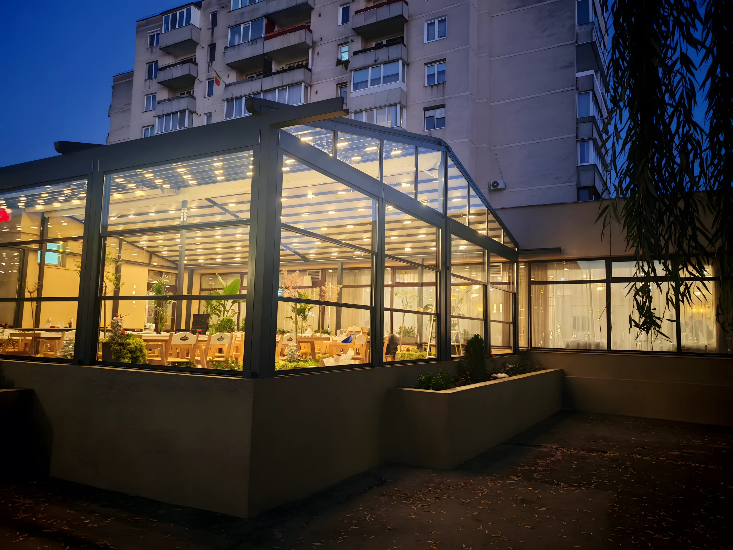 Terasă de restaurant închisă cu structură de aluminiu și vitraj, iluminată cald la ora albastră