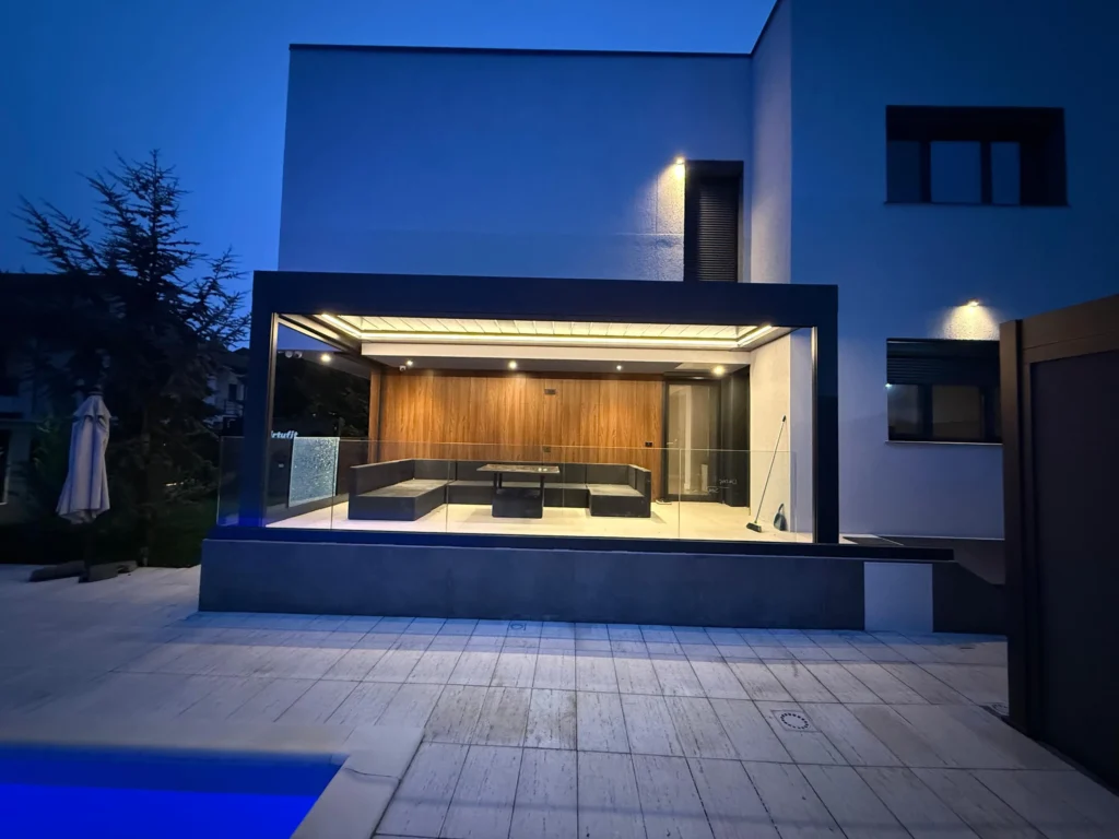 Terasă acoperită cu structură de aluminiu antracit și balustradă din sticlă, iluminată cu LED cald la ora albastră