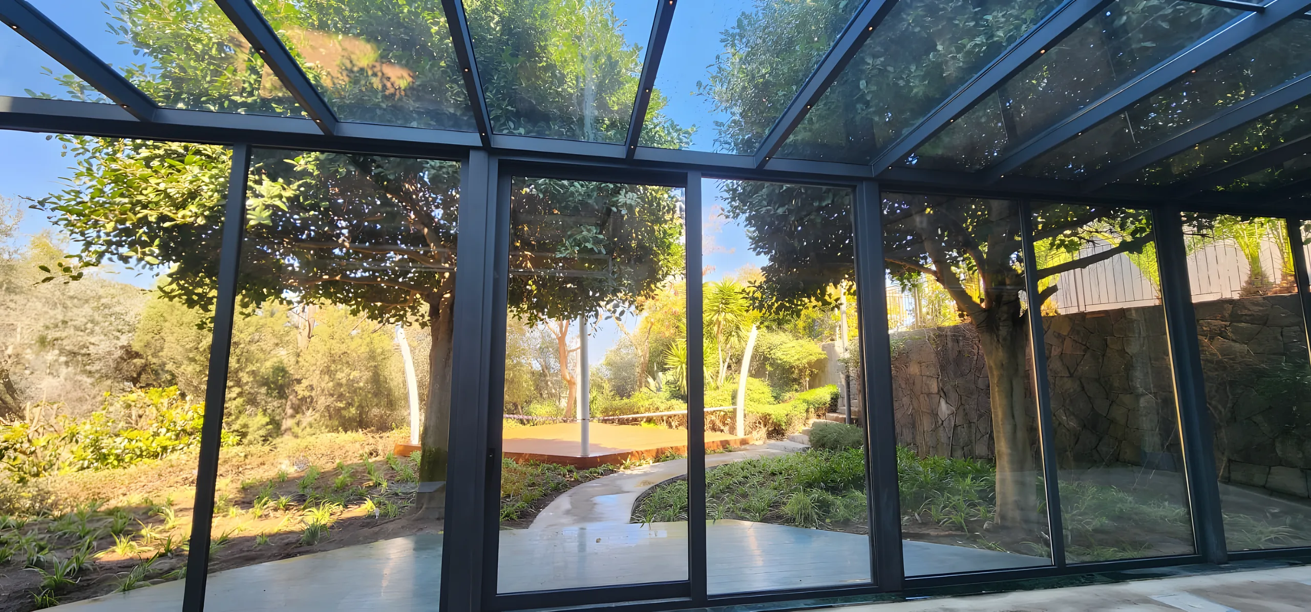 Vedere din interiorul unei verande din aluminiu și sticlă către o grădină cu arbori maturi