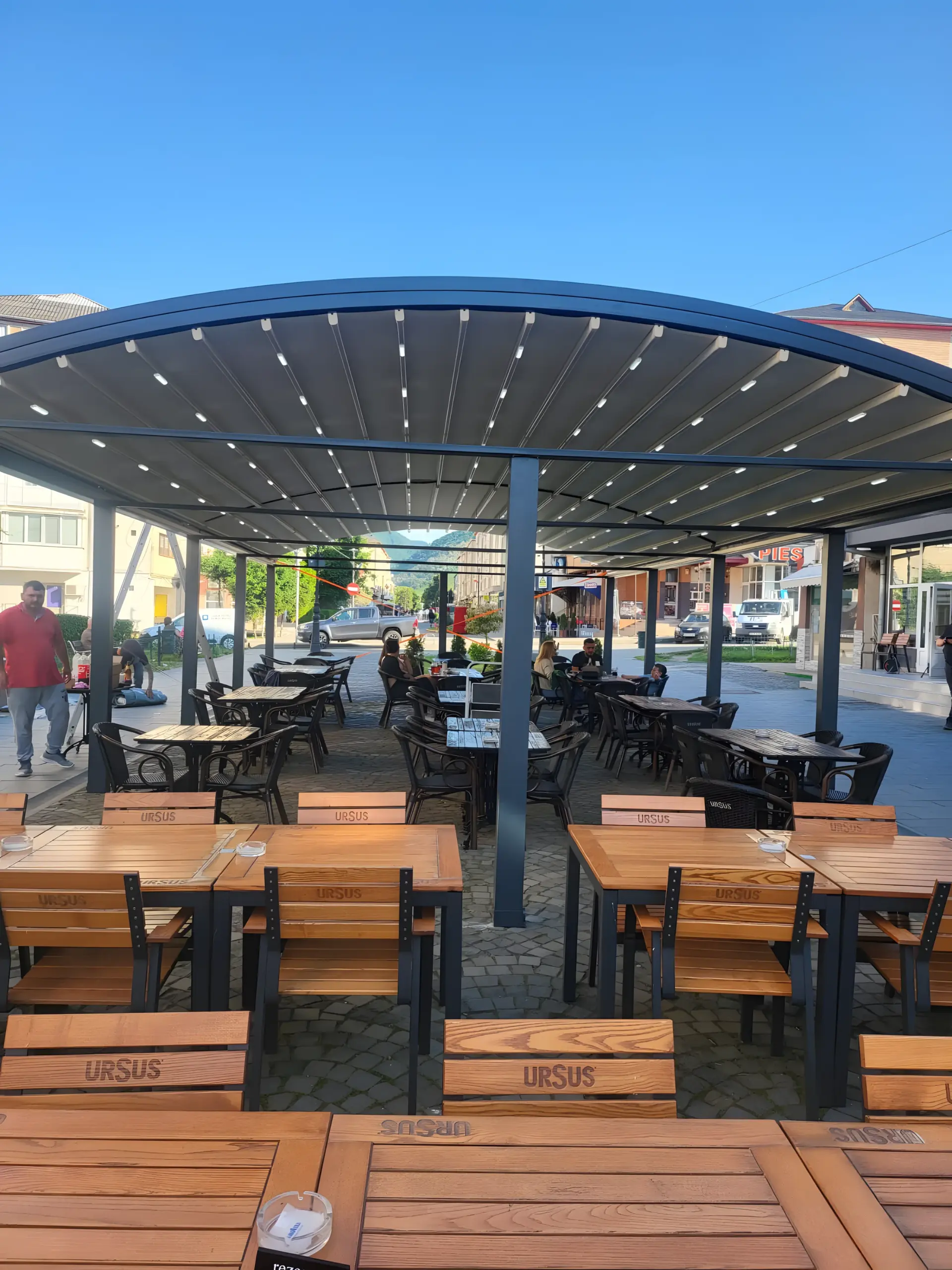 Terasă de local acoperită cu copertină retractabilă pe structură curbată de aluminiu, cu iluminare LED în grinzi