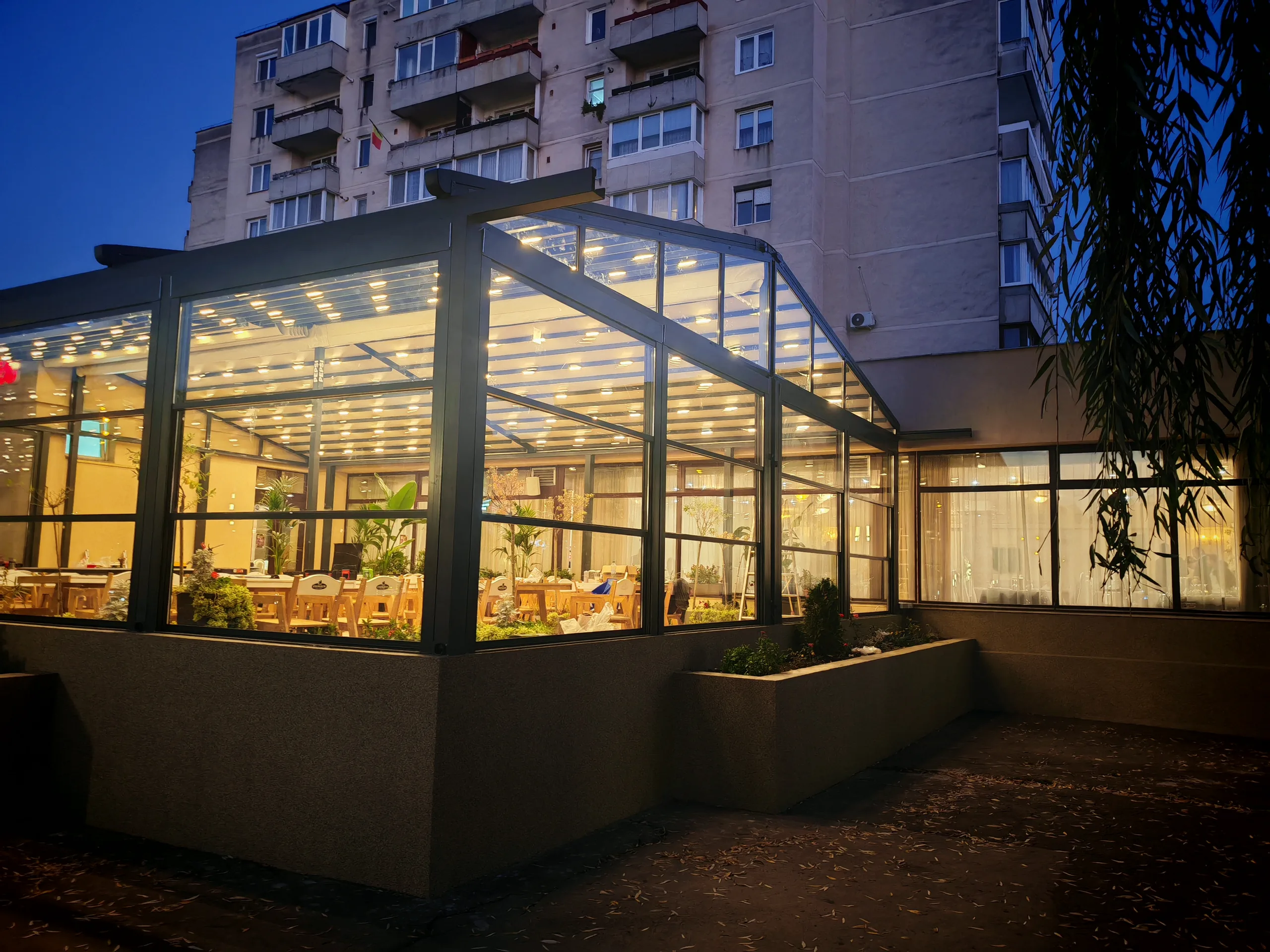 Verandă vitrată din aluminiu antracit, iluminată la ora albastră, cu sală de evenimente vizibilă în interior