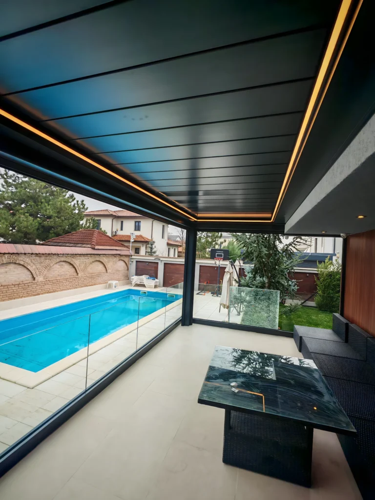 Pergolă bioclimatică cu lame de aluminiu antracit și iluminat LED liniar, deschisă spre piscină cu balustradă din sticlă